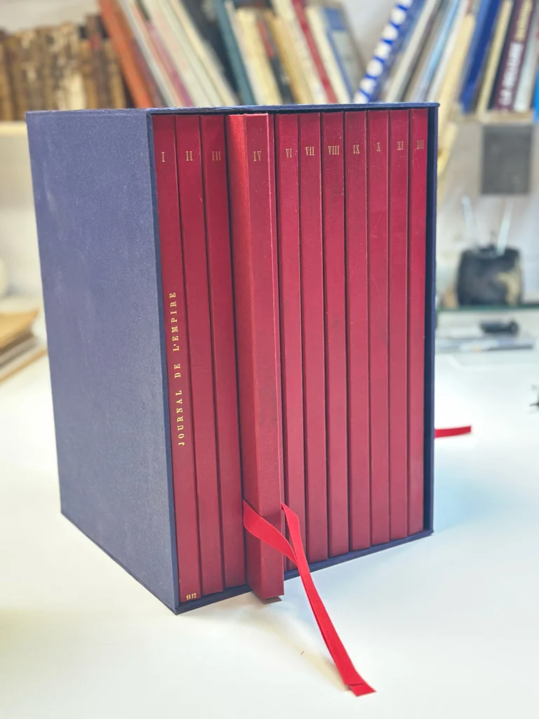 Série de volumes rouges réunis dans un étui de conservation bleu, avec un ruban de préhension.