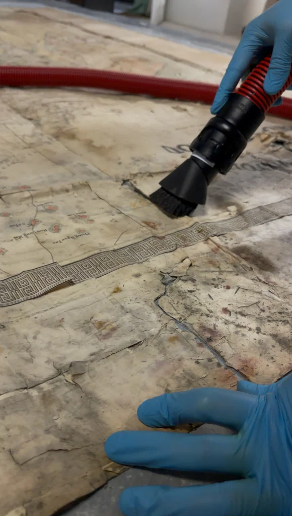 Micro-aspiration d’une carte ancienne à l’aide d’une brosse, avec maintien du document par une main gantée.