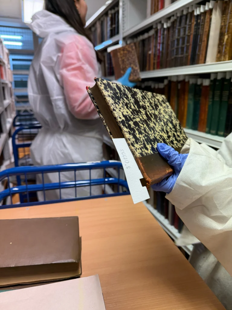 Manipulation d’un volume à couverture marbrée avec des gants et une tenue de protection, devant les rayonnages.