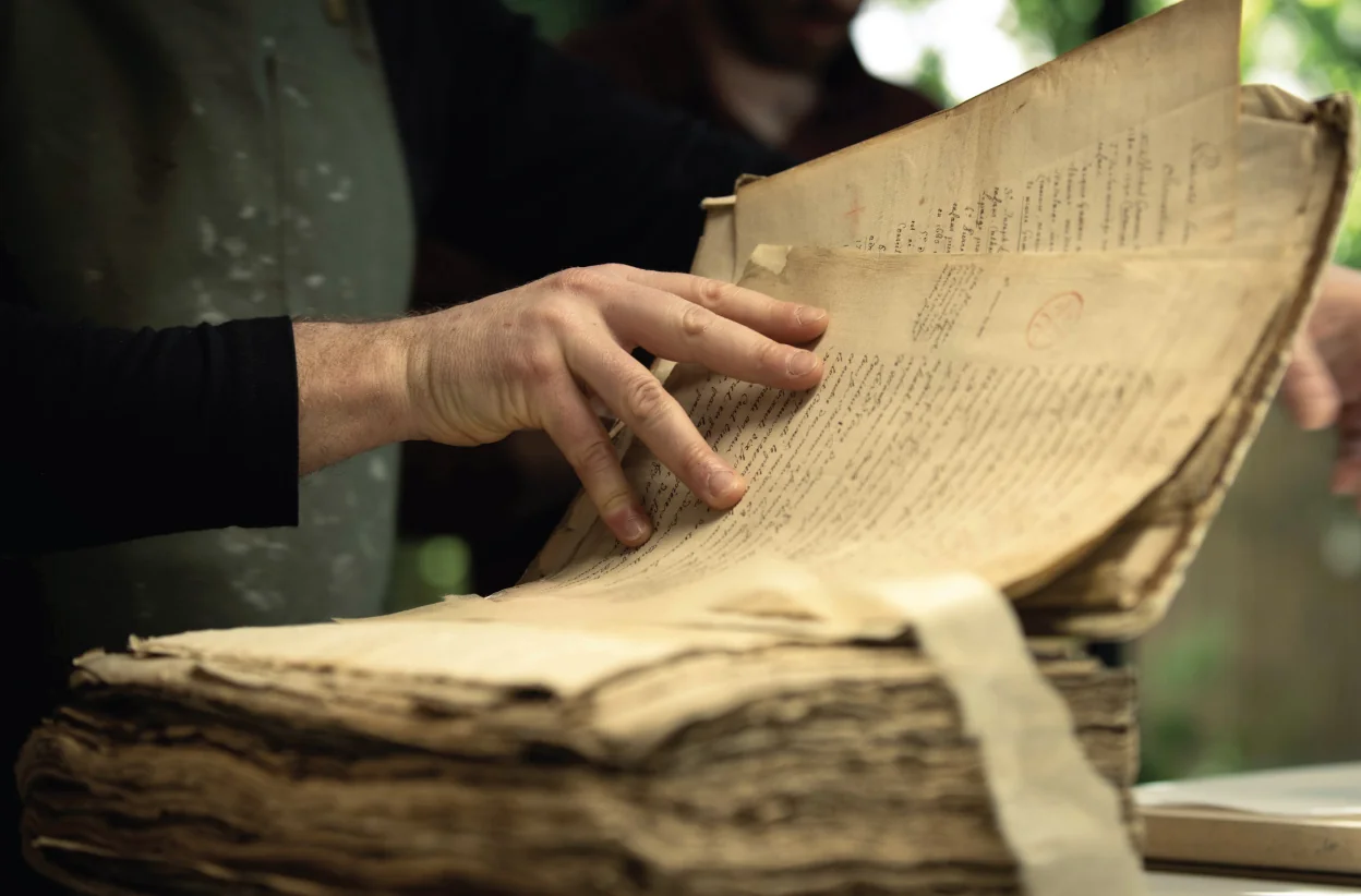 Mains soulevant un feuillet manuscrit dans un ouvrage ancien ouvert.