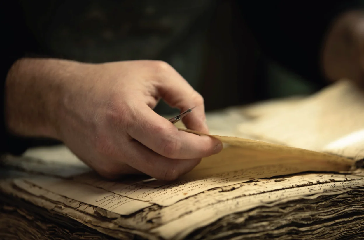 Main tenant une aiguille au-dessus des feuillets d’un ouvrage manuscrit.