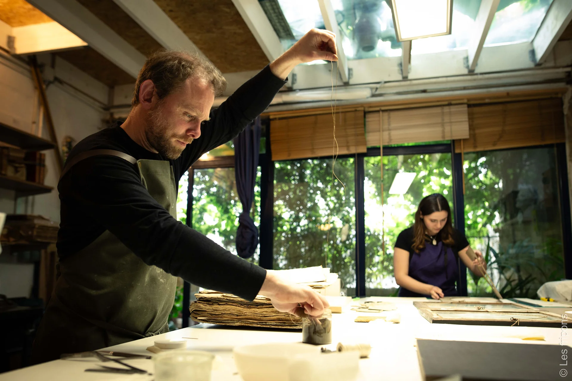 Un artisan travaille sur un ouvrage ancien à l’établi, une seconde personne à l’arrière-plan.
